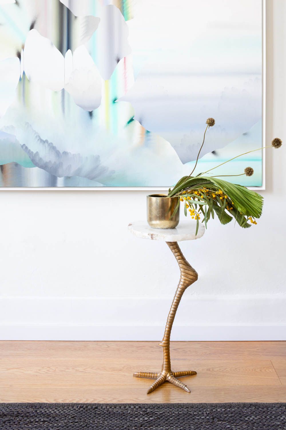 Brass bird-leg side table holding mimosa and a palm frond beneath the artwork