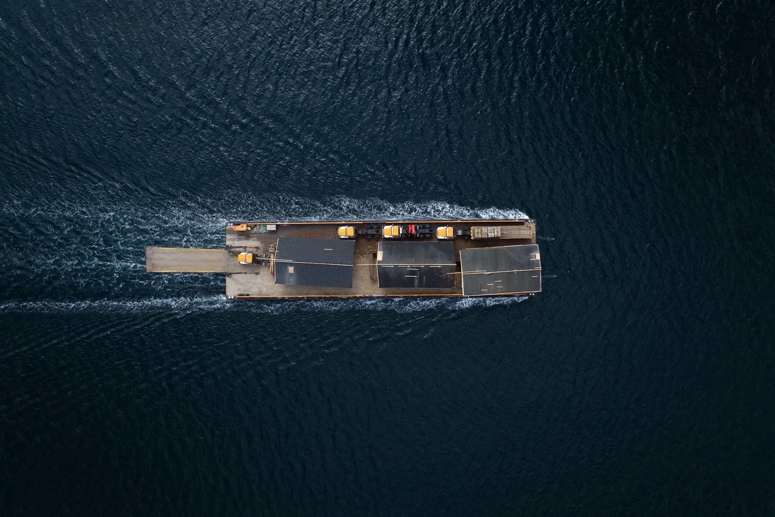 Aerial view of a barge carrying three relocated houses and haul trucks across open water