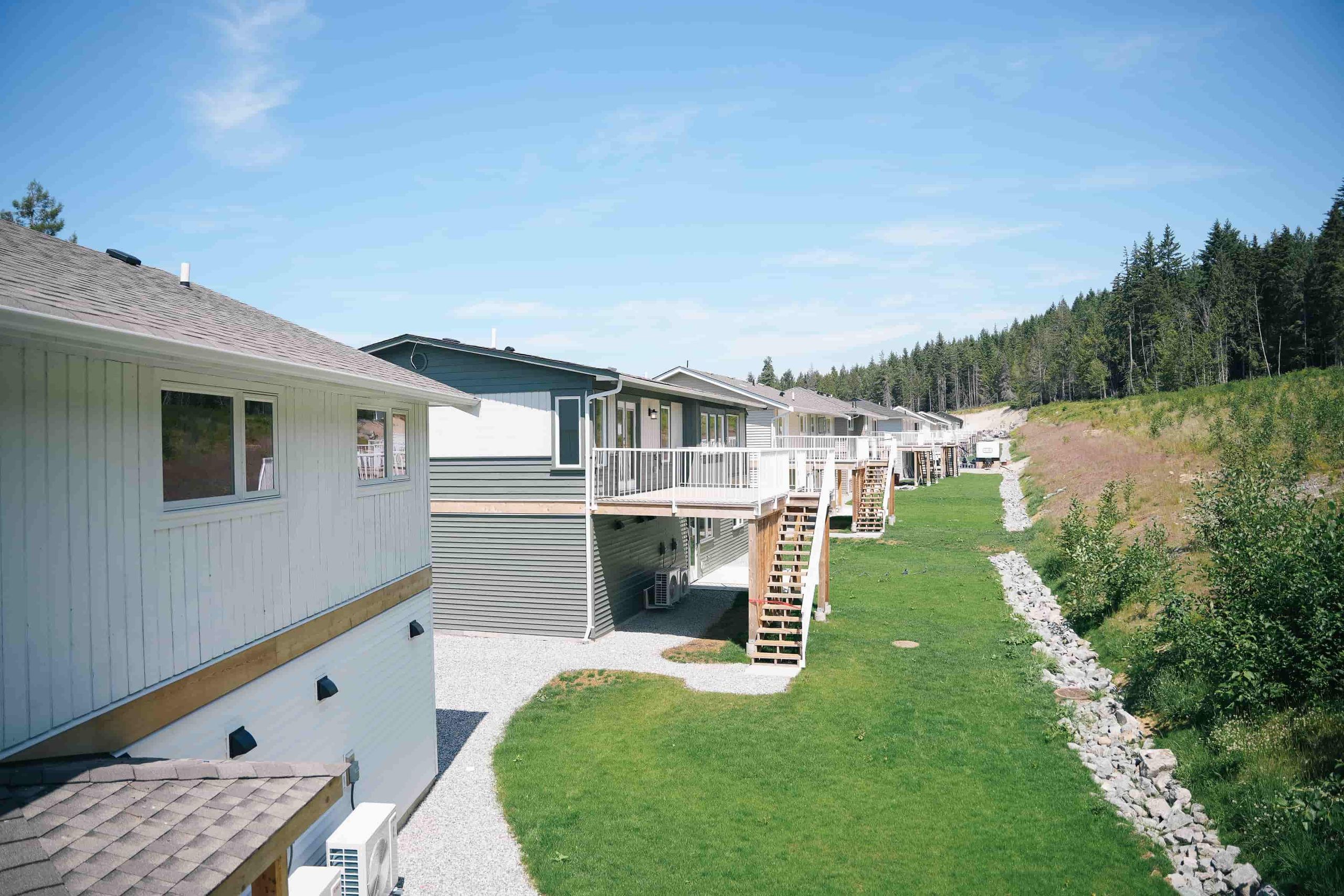 Row of completed homes with new decks and stairs above garden-suite levels, backing onto forest