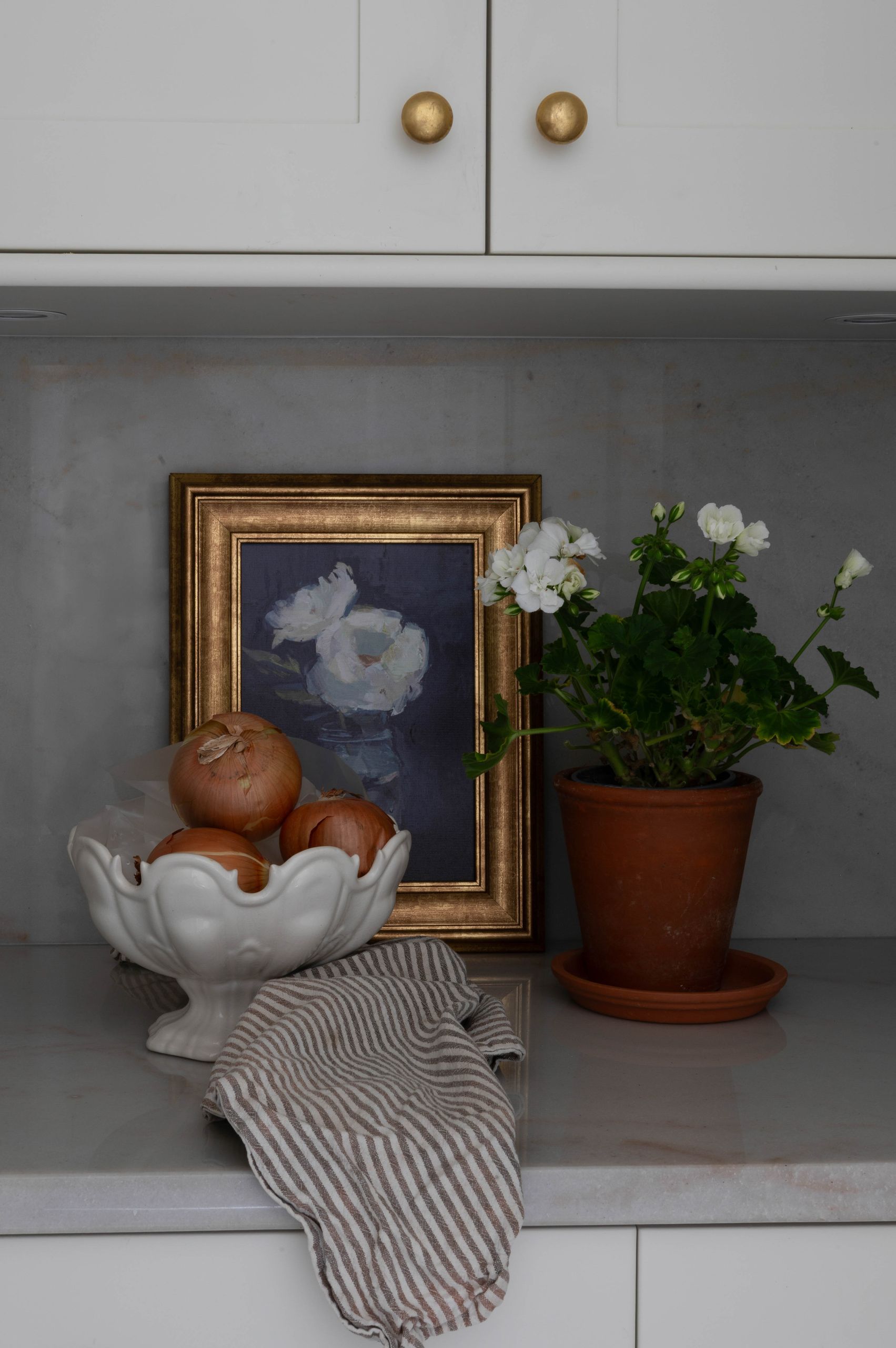 Counter vignette with a gilt-framed still life, a white compote of onions and potted geraniums